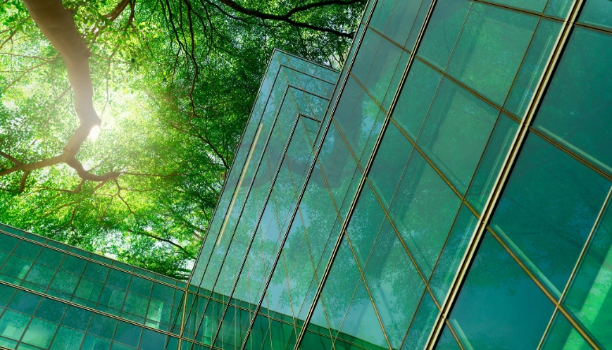 Eco-friendly building in the modern city. Green tree branches with leaves and sustainable glass building for reducing heat and carbon dioxide. Office building with green environment. Go green concept.